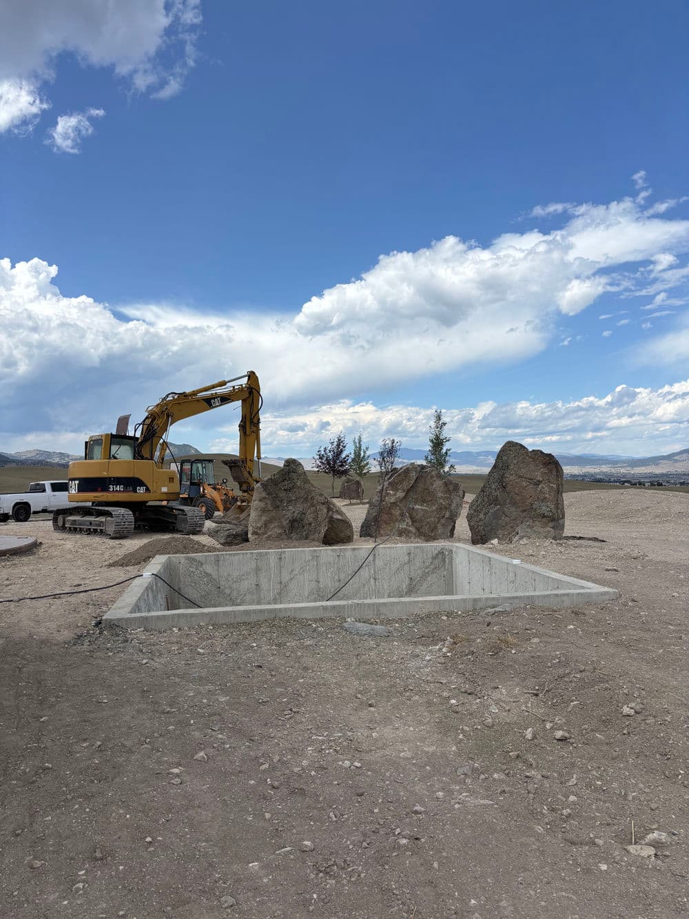 Excavator near large rocks and a concrete foundation in an outdoor construction site.