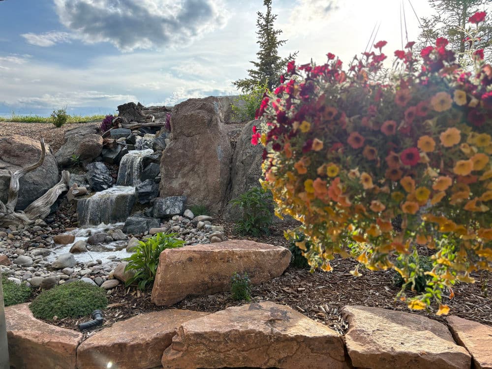 Picturesque waterfall surrounded by colorful flowers and rocky landscape in a serene setting.