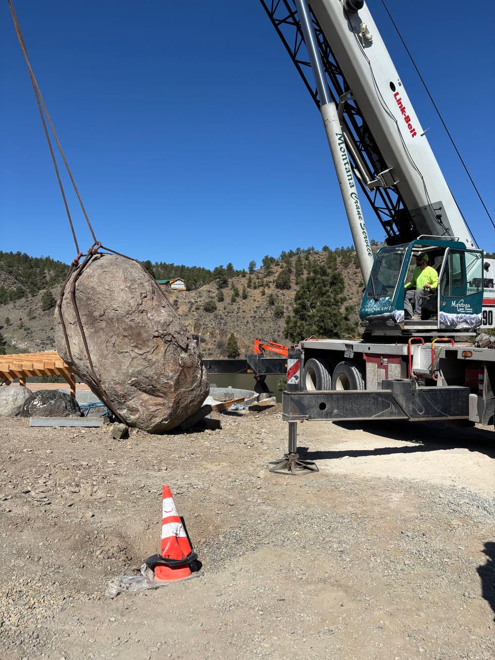 Crane lifting large rock at construction site with orange traffic cone in foreground.