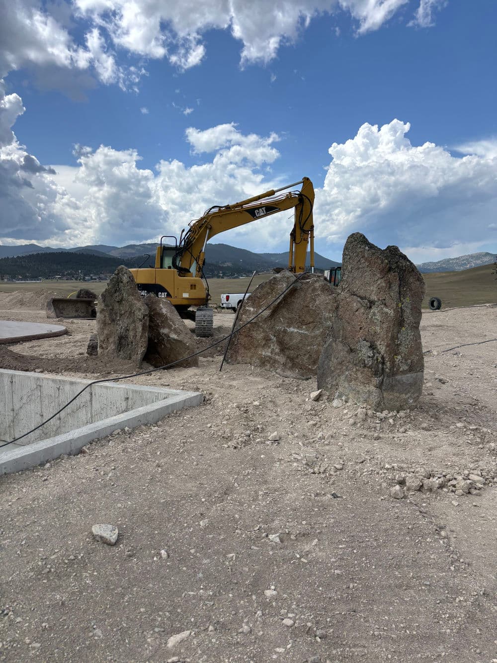 Excavator working on rocky terrain with large boulders under a cloudy sky.