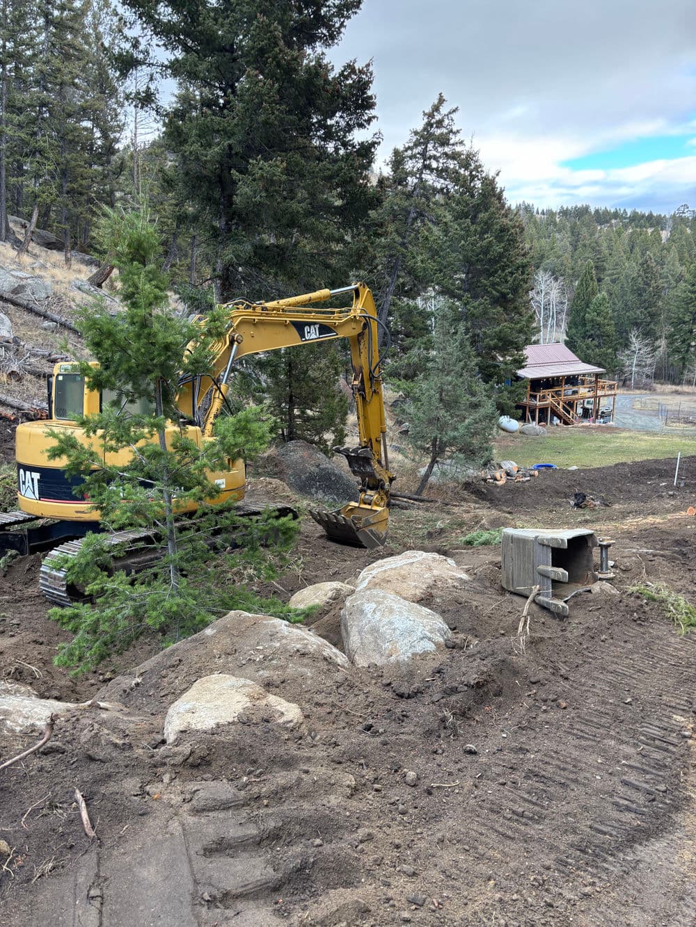 Excavator working on landscape with rocks and trees at a construction site in a rural area.