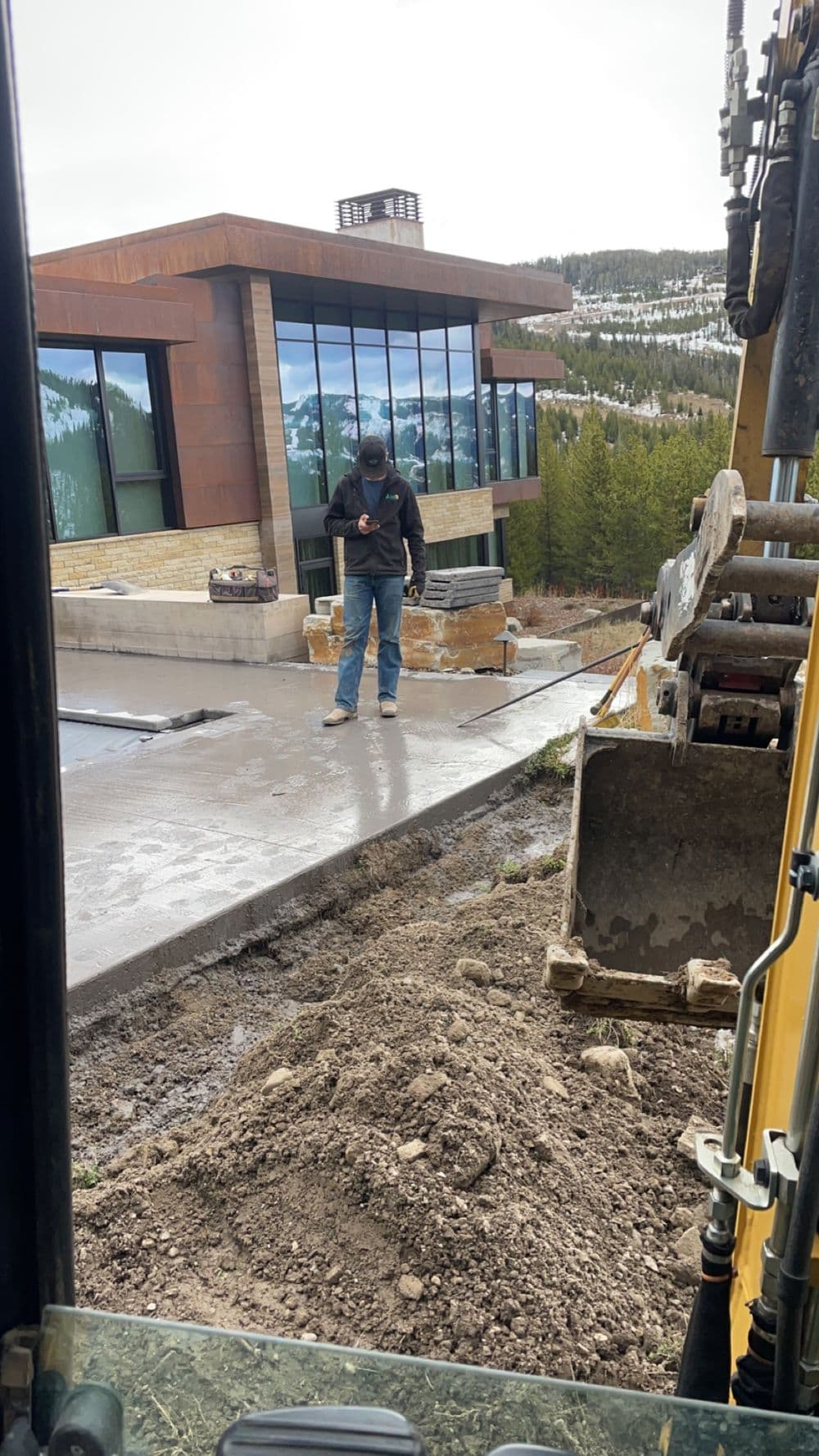 Construction worker using a smartphone near a modern mountain home with panoramic windows.