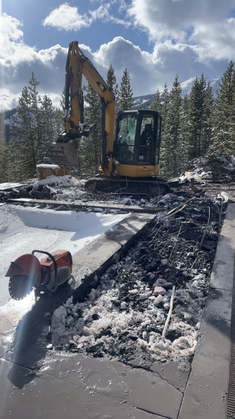 Excavator working on snowy construction site surrounded by pine trees and cloudy sky.