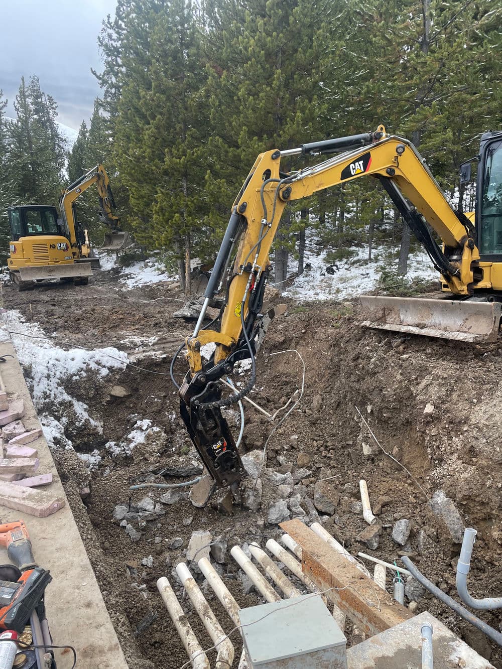 Excavators digging in a forested area, with snow and utility pipes visible.
