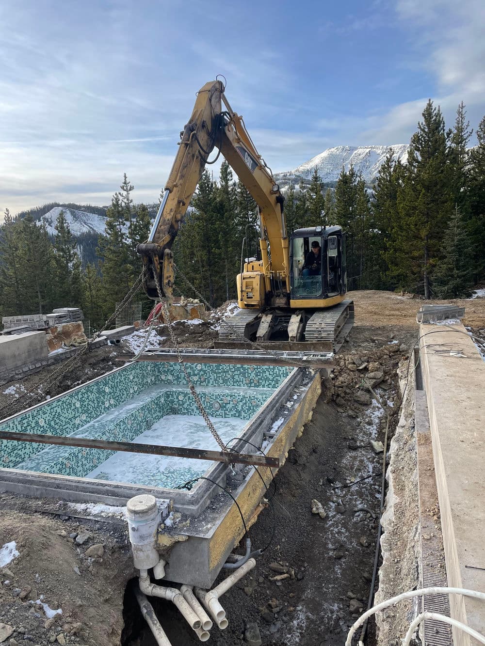 Excavator lifting a swimming pool in a mountainous area with trees in the background.