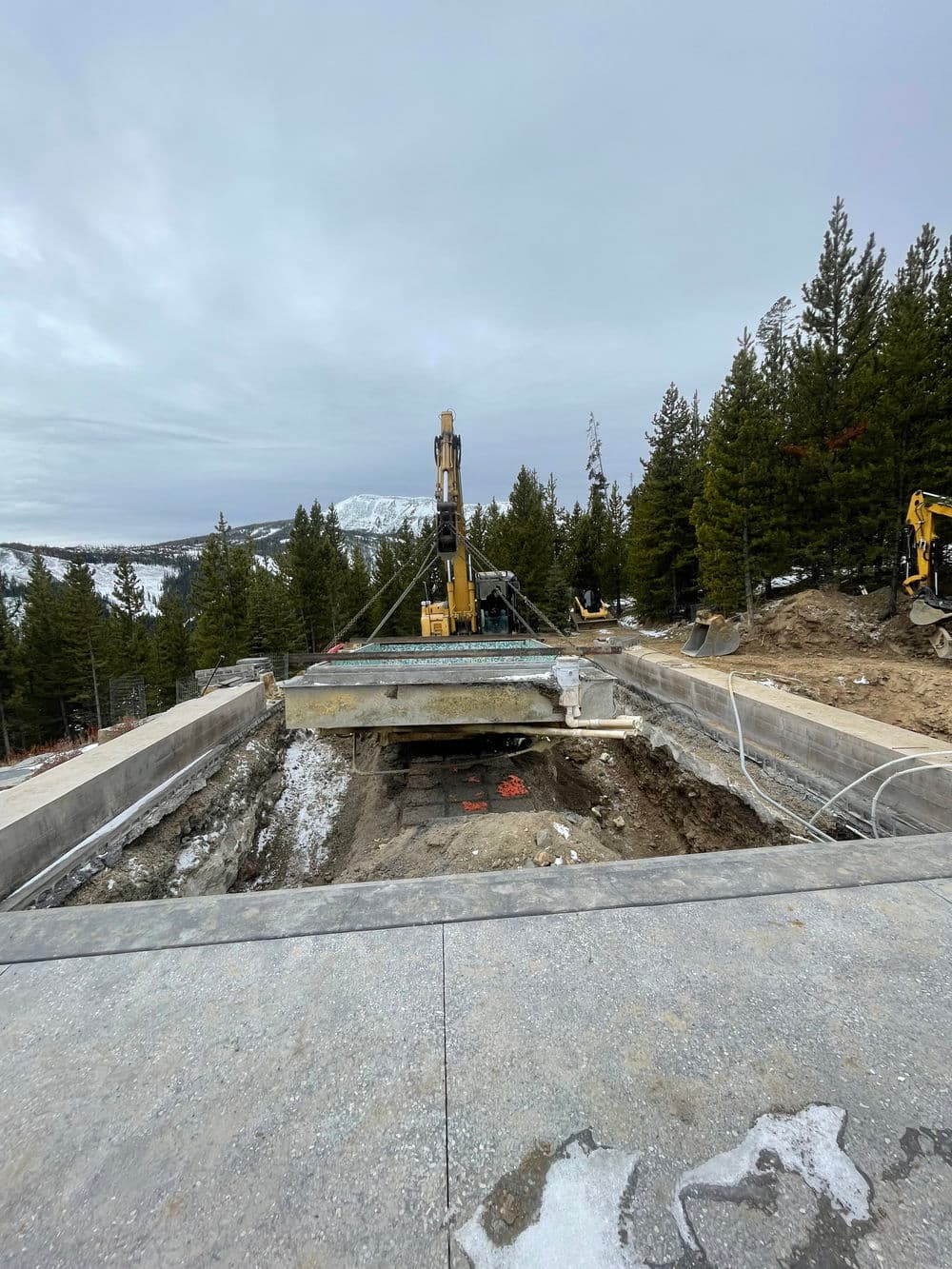 Construction site with heavy machinery and a concrete slab, surrounded by pine trees and mountains.