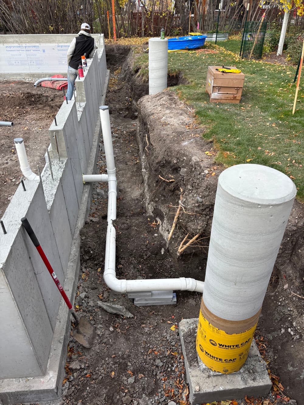 Construction site showing underground plumbing installation with concrete pillars and trench.