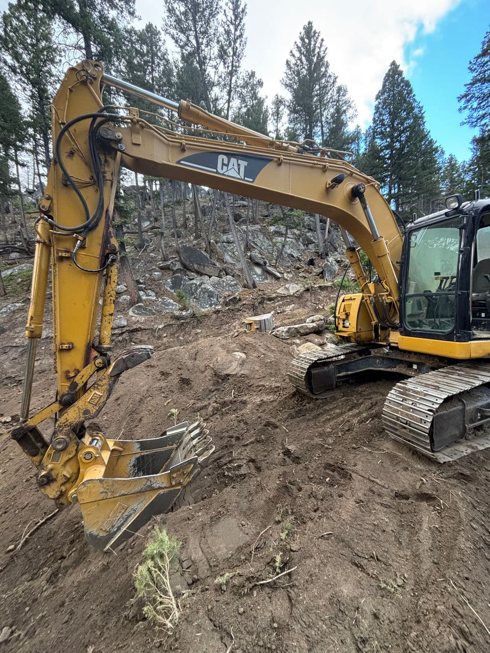 CAT excavator on a construction site, surrounded by trees and rocky terrain.
