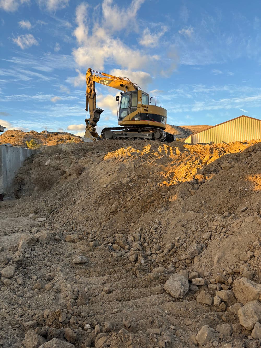 Caterpillar excavator on a dirt mound under a blue sky with scattered clouds.