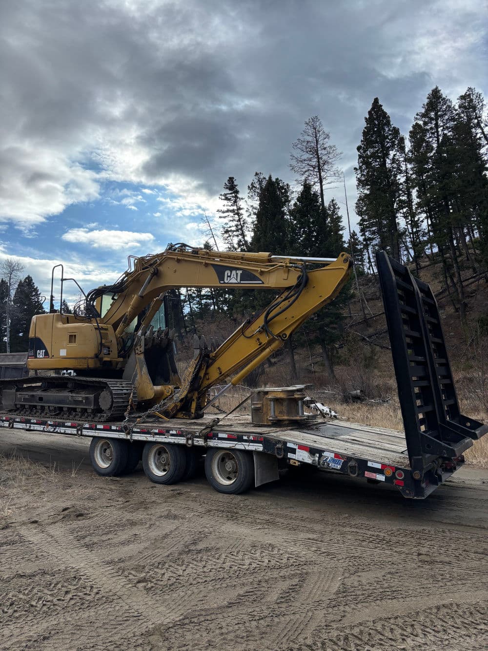 CAT excavator loaded on a flatbed trailer in a rugged outdoor setting with pine trees.